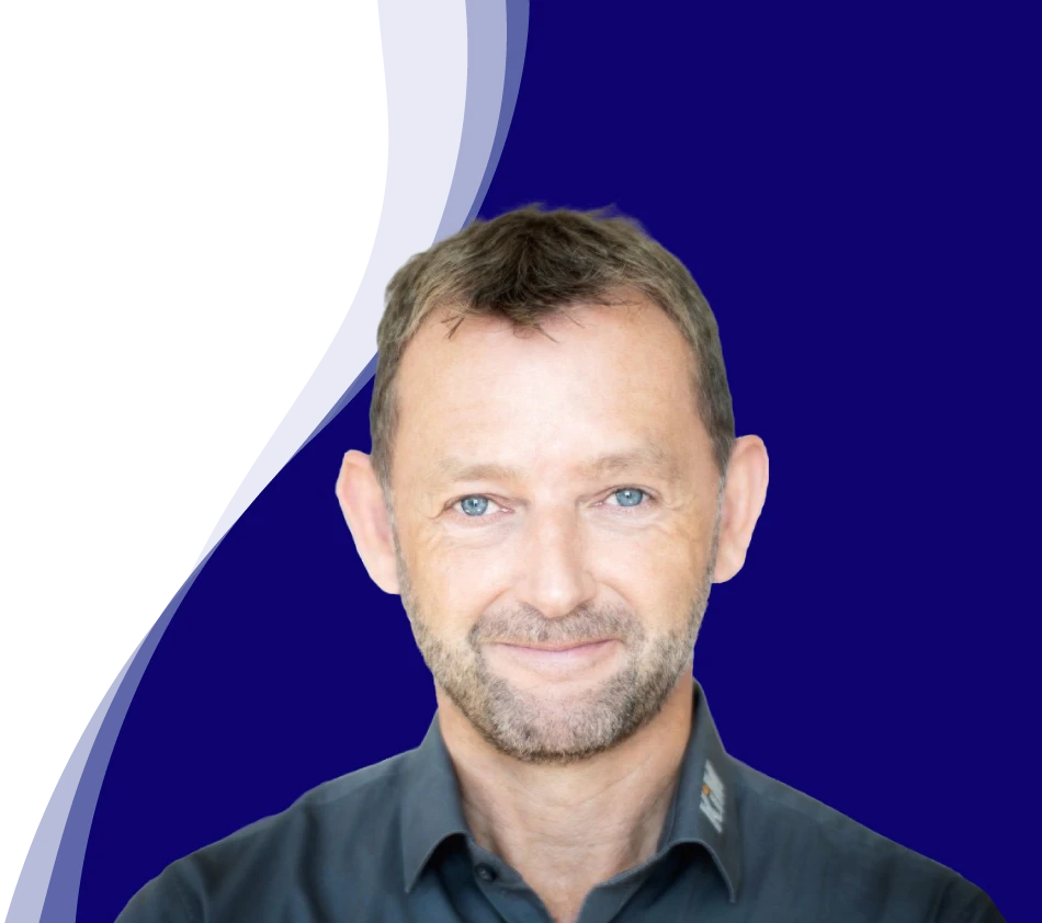 Achim Angel, directeur de KiM GmbH - Member of 4PACE, sourit aimablement à la caméra. Il a les cheveux bruns courts, une courte barbe et porte une chemise gris foncé avec un col légèrement ouvert. En arrière-plan, une forme ondulée incurvée en blanc, bleu clair et bleu foncé confère à la photo un design moderne.